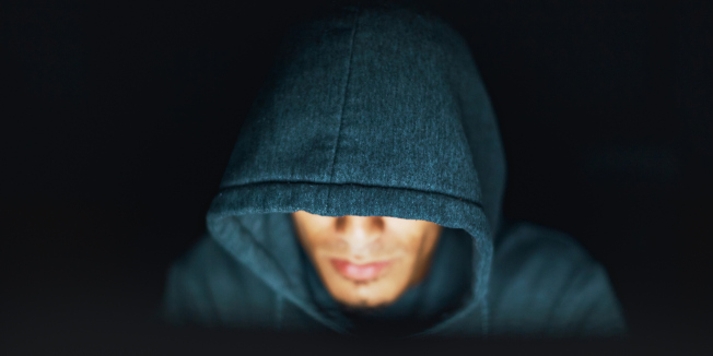 A young man, face partly obscured by a hooded shirt, works on an unseen but illuminated computer by night.