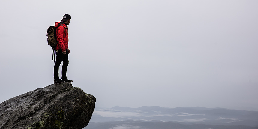 Junger Mann steht auf einem großen Felsblock mit Blick auf ein Gebirge