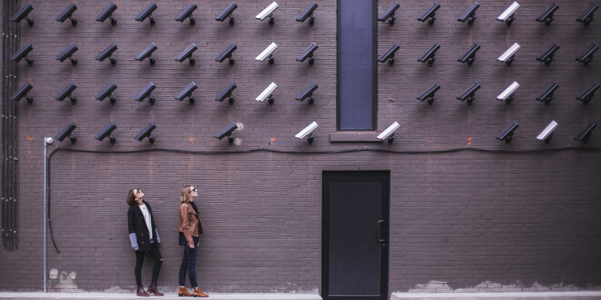 Two women looking up at a brick wall covered in surveillance cameras.