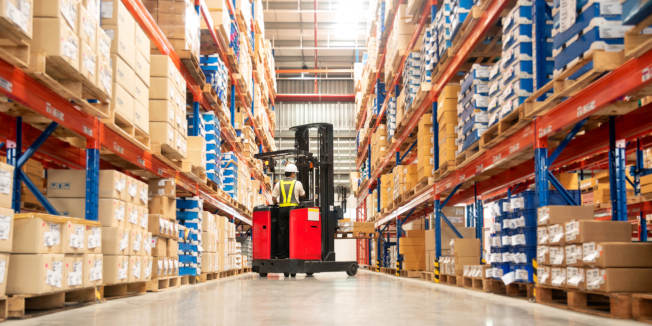 Worker in forklift-truck loading packed goods in huge distribution warehouse with high shelves.