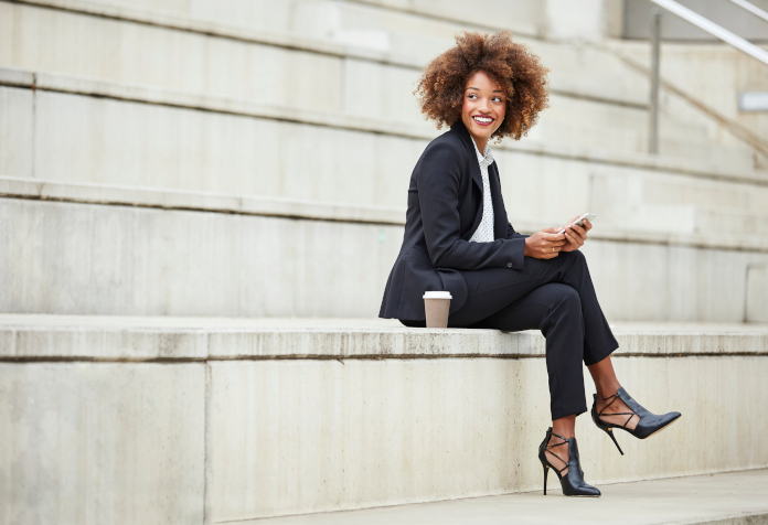 Happy businesswoman looking away while holding smart phone on steps. Female professional is sitting with legs crossed and knee on staircase. She is in formals.