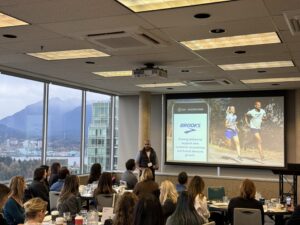 A conference room with a presentation on "Brooks" discussing customer acquisition and brand growth, featuring an image of two people running. Attendees are seated, with windows showing a mountain and cityscape view.