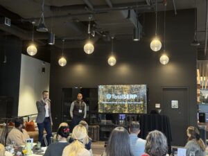 People attending a presentation with a speaker and a screen displaying "TotalSERP" in a dimly lit room.