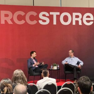 Two people seated on stage with "RCCSTORE" backdrop, engaged in discussion with an audience watching.