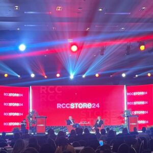 Two people sitting on stage with "RCC STORE 24" displayed on large red screens, surrounded by an audience and colourful stage lights.