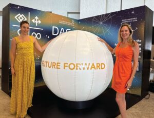 Two women standing beside a large white sphere with the text "FUTURE FORWARD" in an exhibition area.