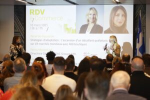 A crowded conference room with two women speaking on stage in front of a large presentation screen displaying details of an e-commerce panel discussion.