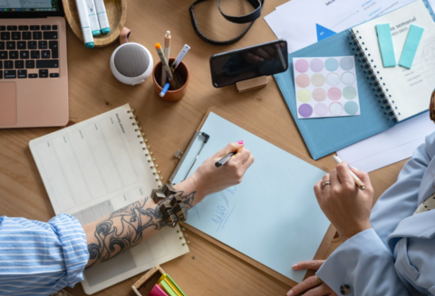 Two people collaborating at a desk with a laptop, notepads, and markers.