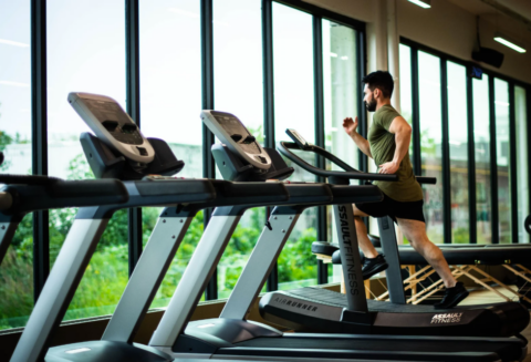 A man running on a treadmill in a gym with large windows.