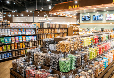 Grocery store interior with shelves of bulk candies in transparent containers, a sushi station visible in the background.