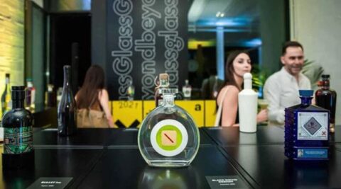 Dos personas de pie detrás de una mesa con varias botellas de licor exhibidas en un ambiente moderno e iluminado.