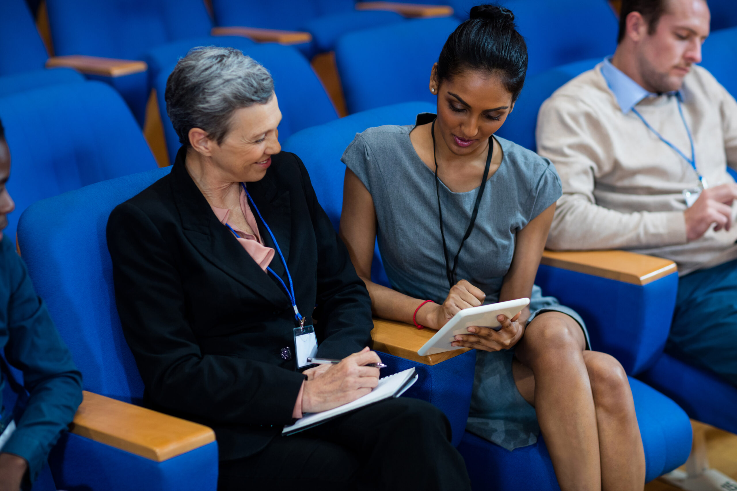 Two people sitting on blue chairs, one using a tablet and the other taking notes, with another person in the background.