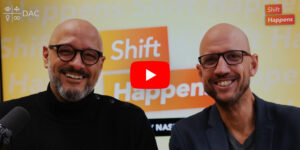 Two men smiling in front of a "Shift Happens" background with a video play button overlay.