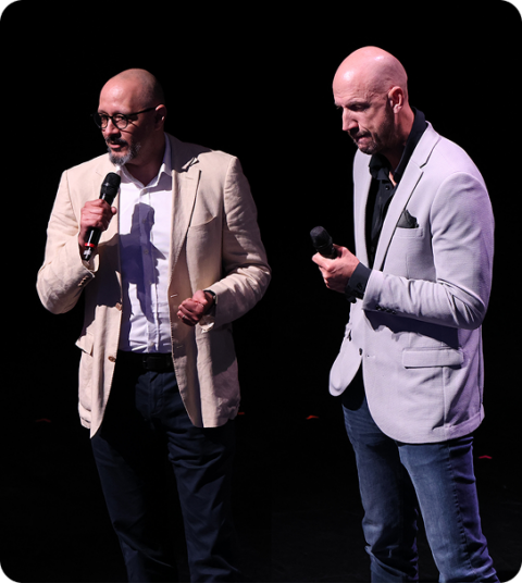 Two men wearing blazers stand on stage holding microphones, speaking to an audience.