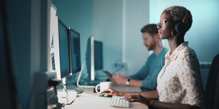 Closeup side view of mid 20's blond black woman doing her design project on a computer. She might be a software developer as well. She's sitting in front of a desktop computer and sipping a coffee while working on a computer.